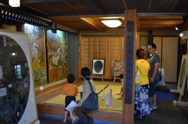 畳の部屋に大きな絵が飾られていてそれを子供や大人たちが見ている写真